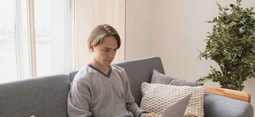 Young white man sitting on a couch cross legged working on a laptop
