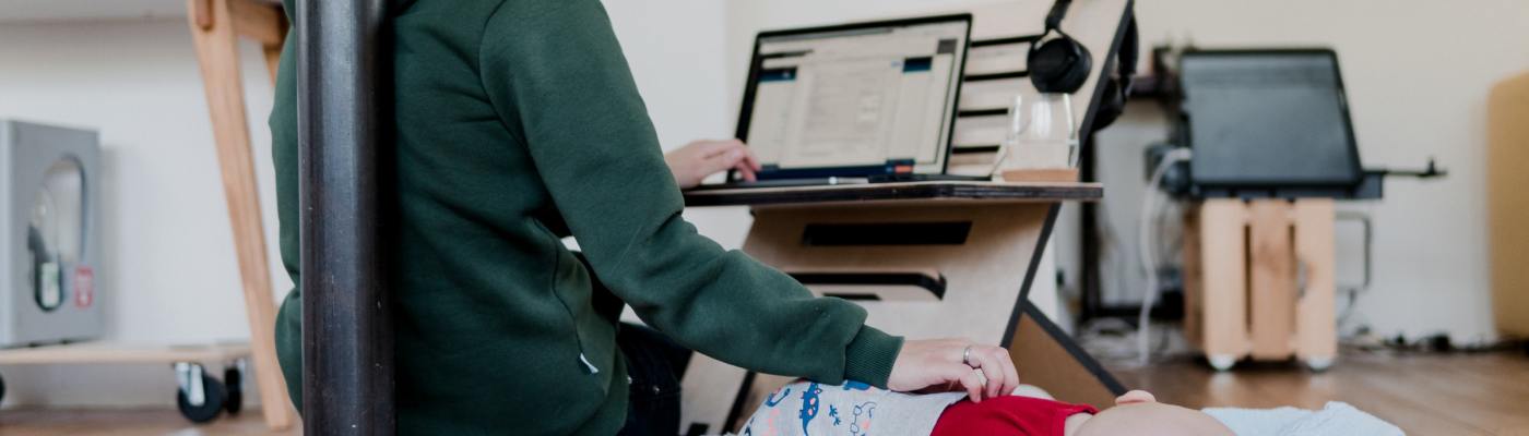 Person working on laptop while tending to baby on a mat on the floor.