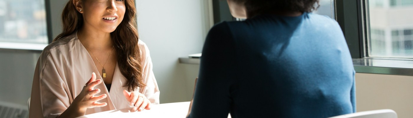 Two people sitting at a table talking; one's face is in view and the other's back is in the frame